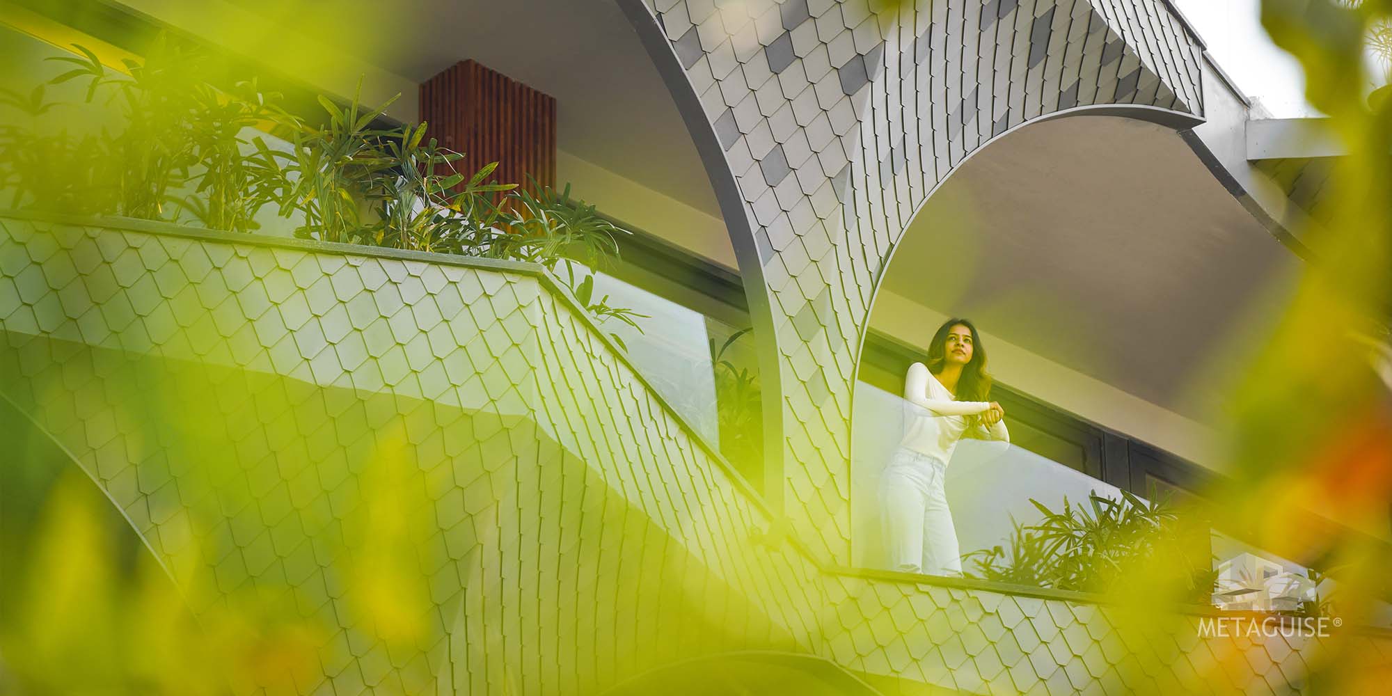 Woman standing on a balcony framed by an arched metal facade clad in scale-pattern panels