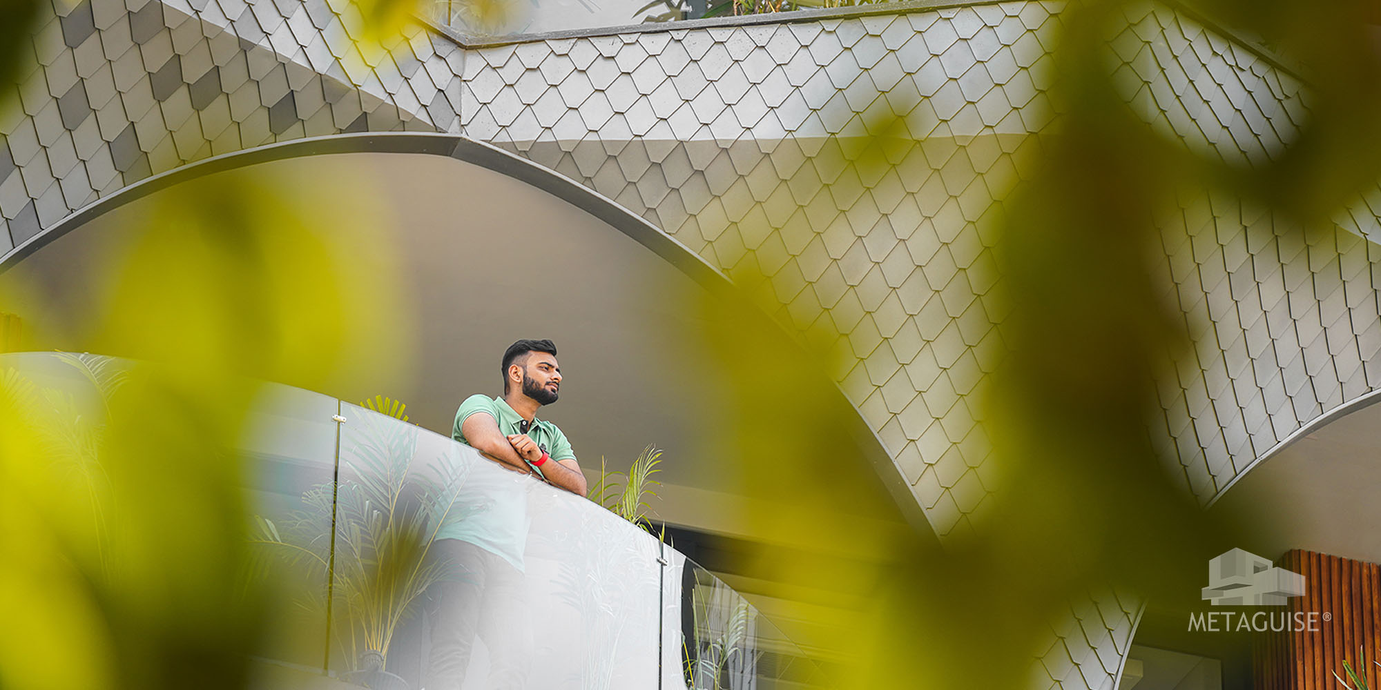 Man looking out from a balcony framed by overlapping scale-pattern aluminium cladding and green leaves