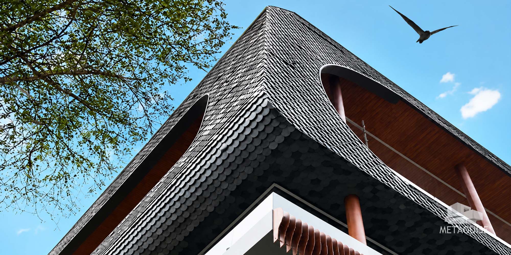 Upward view of a building corner clad in dark metal scale-pattern panels with a bird in the sky