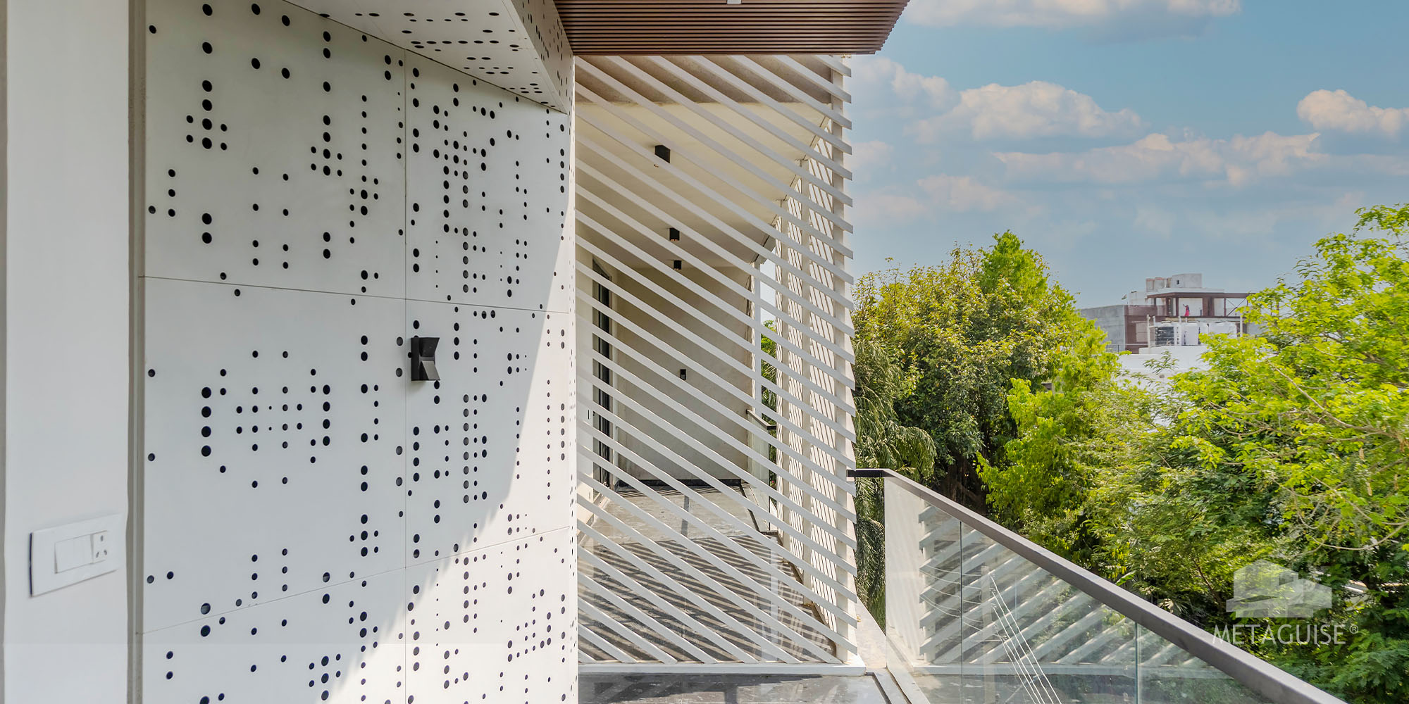 White perforated metal panel wall beside a louvered balcony screen with trees in the background